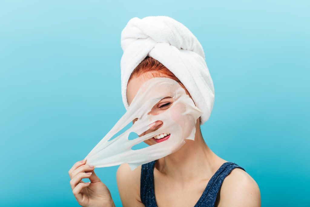 Joyful caucasian girl taking off face mask. Studio shot of carefree young woman doing skincare treatment on blue background.